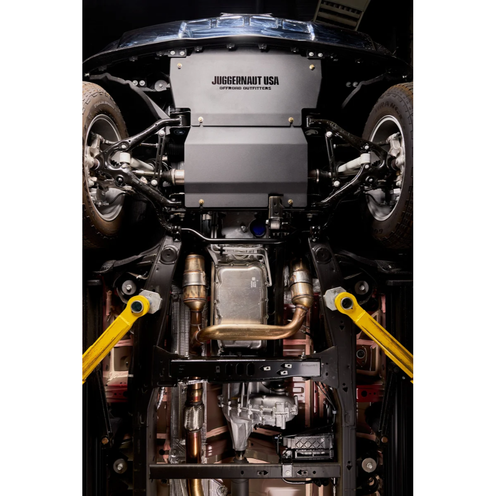 Undercarriage view of a vehicle showing a large gray JUGGERHUNT USA engine cover, yellow suspension arms, exhaust pipes, and mechanical components.
