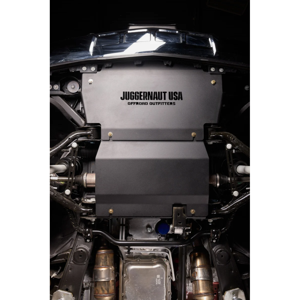 A matte black Juggernaut USA Offroad Outfitters engine cover installed on a vehicle, shown from a top-down view with the engine bay and exhaust components visible.