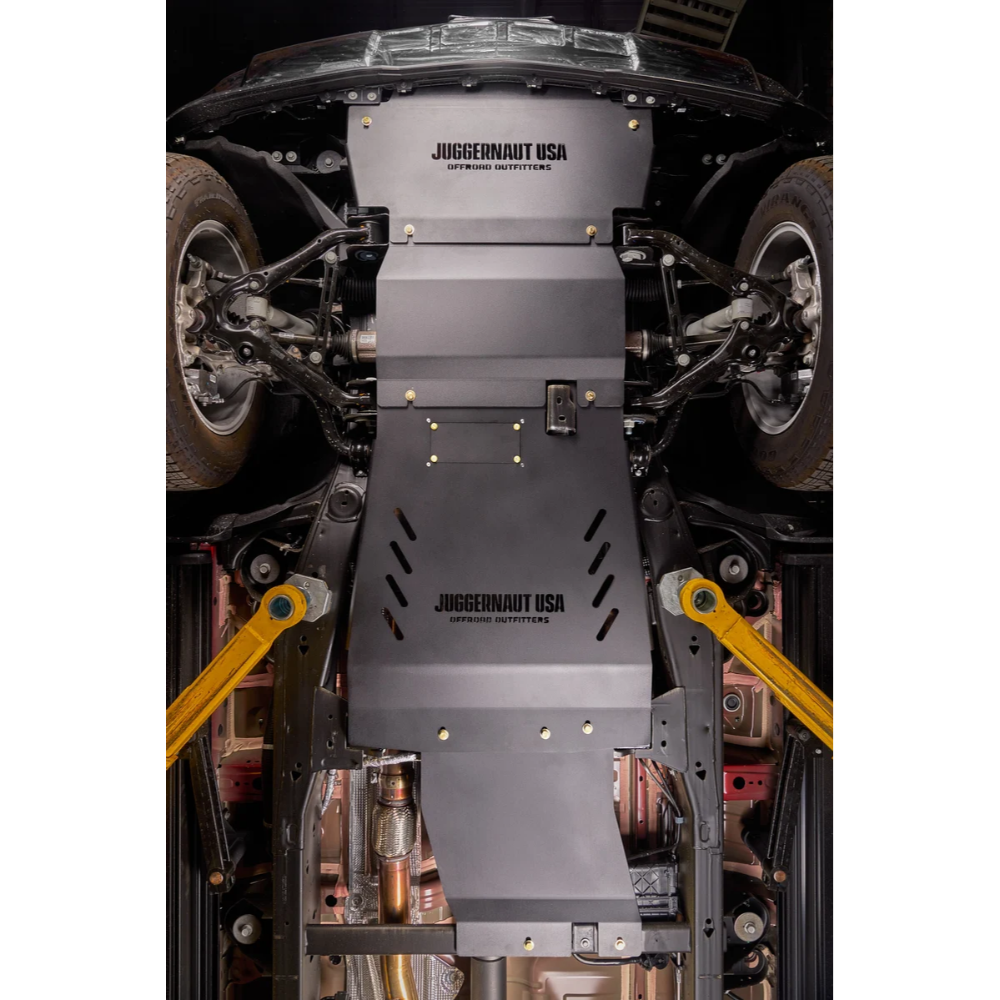 A Juggernaut USA approved steel underbody shield installed on a vehicle chassis, shown from a top-down view with the vehicle's wheels and suspension visible.