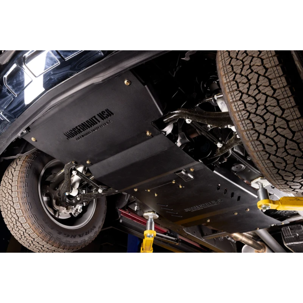 Black steel undercarriage shield and skid plate installed on a vehicle, with visible suspension, tires, and yellow support jacks in a workshop setting.