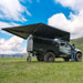 Off-road vehicle with an extended roof awning in a grassy field under a blue sky.
