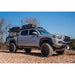 Silver pickup truck with roof racks on a gravel surface with trees in the background