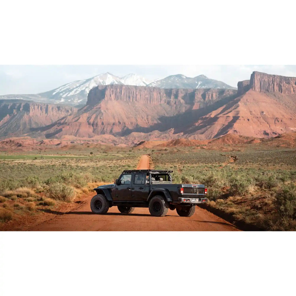 Black off-road gladiator with mounted QDHD Sherpa roof rack on a dirt road with red rock formations and snow-capped mountains in the background.