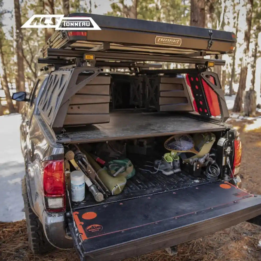 GMC bed with a tonneau cover and ACS Bed Rack, including a cooler and tools, in a forest setting.