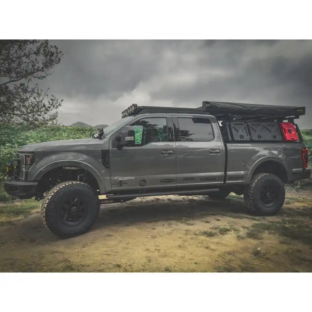 Gray SuperDuty truck with a black roof rack and bed cover on a dirt road with trees and cloudy sky in the background.
