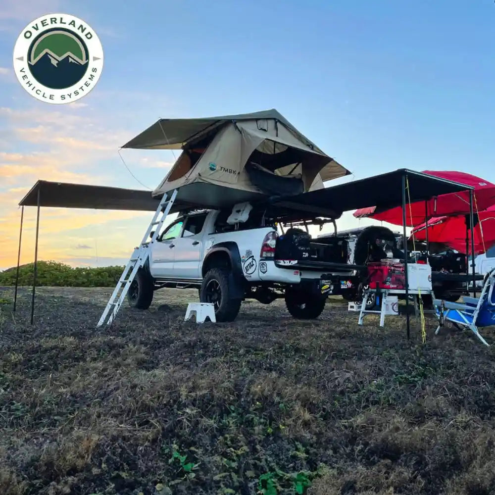 Truck with rooftop tent and overlanding equipment in a field, featuring Overland Vehicle Systems logo.