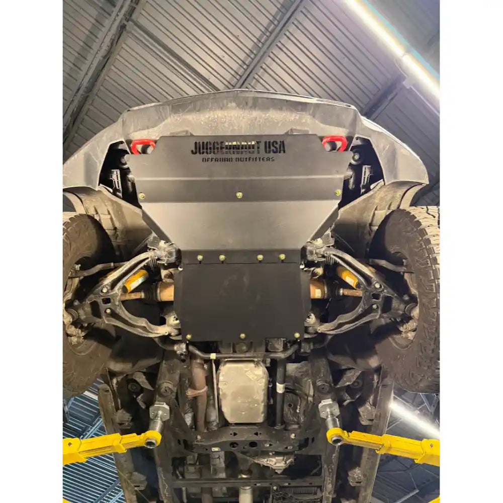 A black metal undercarriage shield and skid plate installed on the rear of a vehicle, photographed from underneath inside a garage. The vehicle is elevated on yellow vehicle lift jacks.