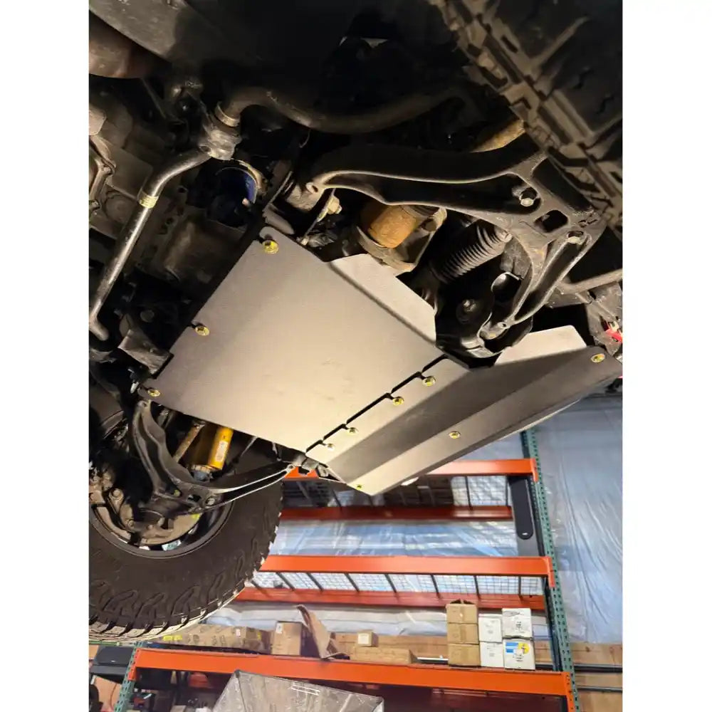 A silver metal undercarriage splash shield with gold-colored screws, installed on a vehicle's chassis in a workshop setting.