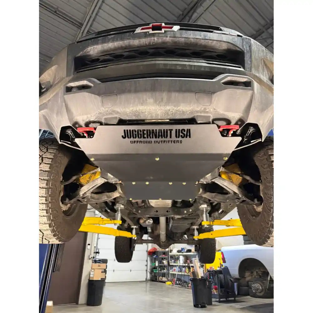 A heavy-duty silver underbody shield with 'JUGGERNAUT USA OFFROAD SUPPITERS' printed on the front, installed on the underside of a lifted black Chevrolet truck on a yellow hydraulic lift inside a garage.
