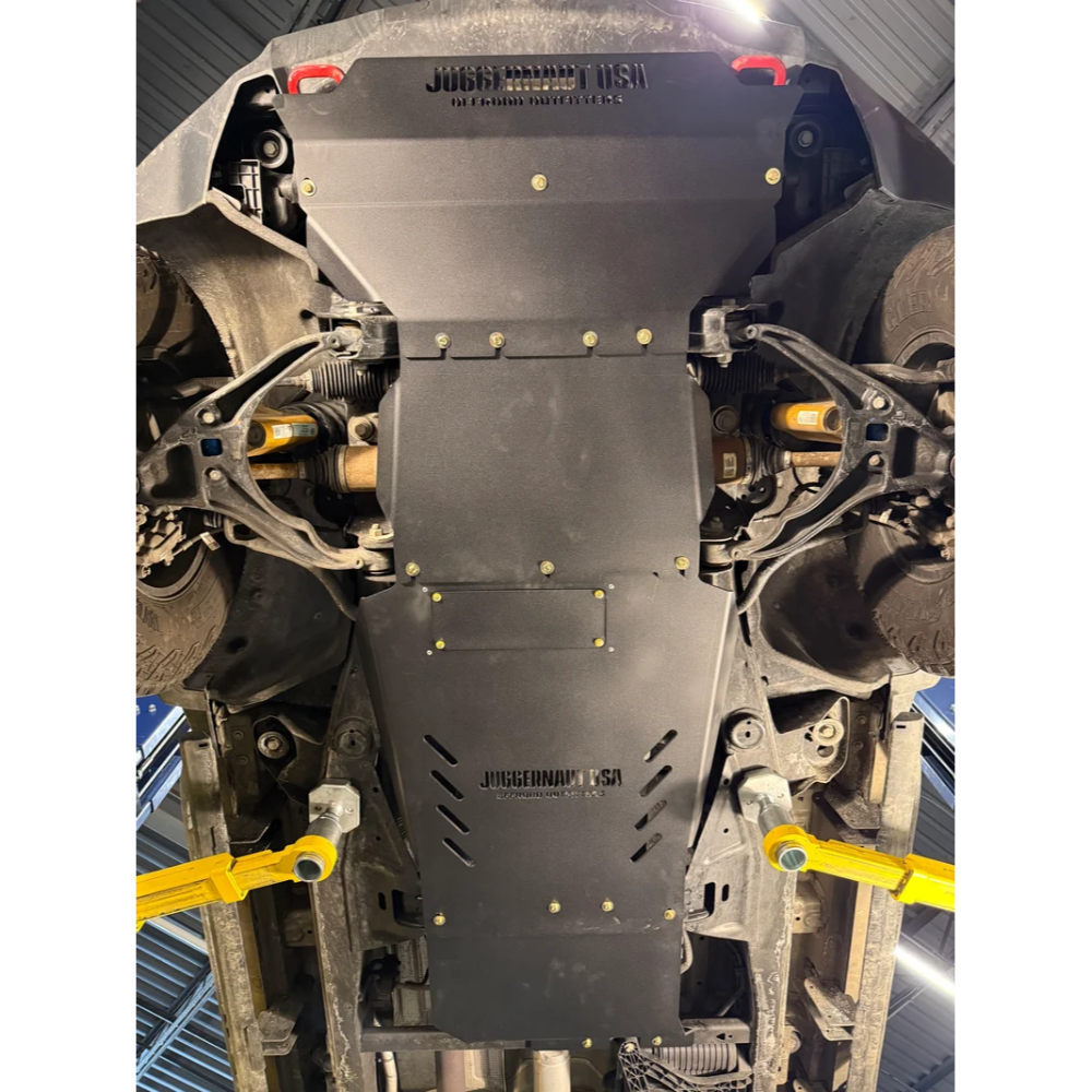 A black steel underbody shield with mounting holes and Juggernaut USA branding, installed on the bottom of a vehicle raised on yellow lifts in a garage.
