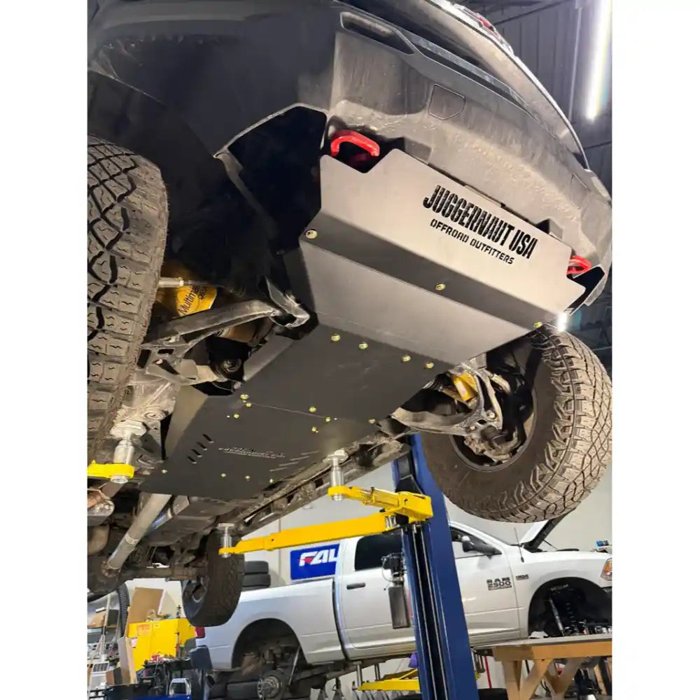 A heavy-duty black underbody skid plate with a silver metal plate reading JUGGERNAUT OFFROAD OUTFITTERS' mounted on the front of a lifted truck undercarriage, shown in an auto repair shop.