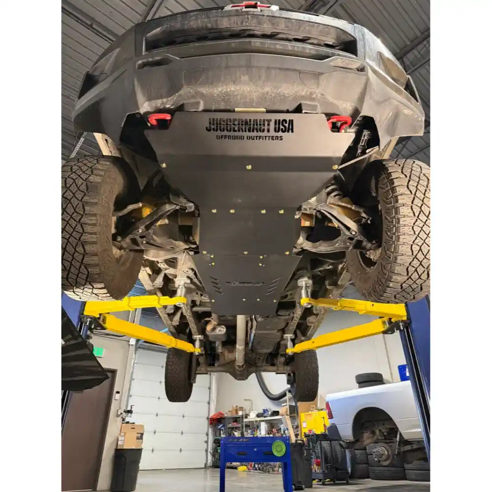 A heavy-duty black underbody shield for a truck, branded 'JUGGERNAUT USA', installed on the bottom of a lifted vehicle in a garage setting.
