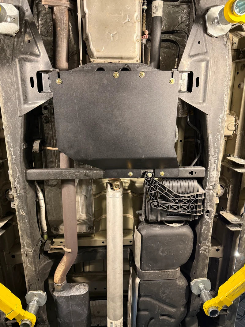 A black metal undercarriage shield mounted on a vehicle's exhaust bracket, shown from underneath on a vehicle frame.