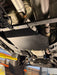 A black metal undercarriage splash shield plate mounted on a vehicle, shown from a low angle inside a garage or workshop with a tire, suspension components, and exhaust visible.