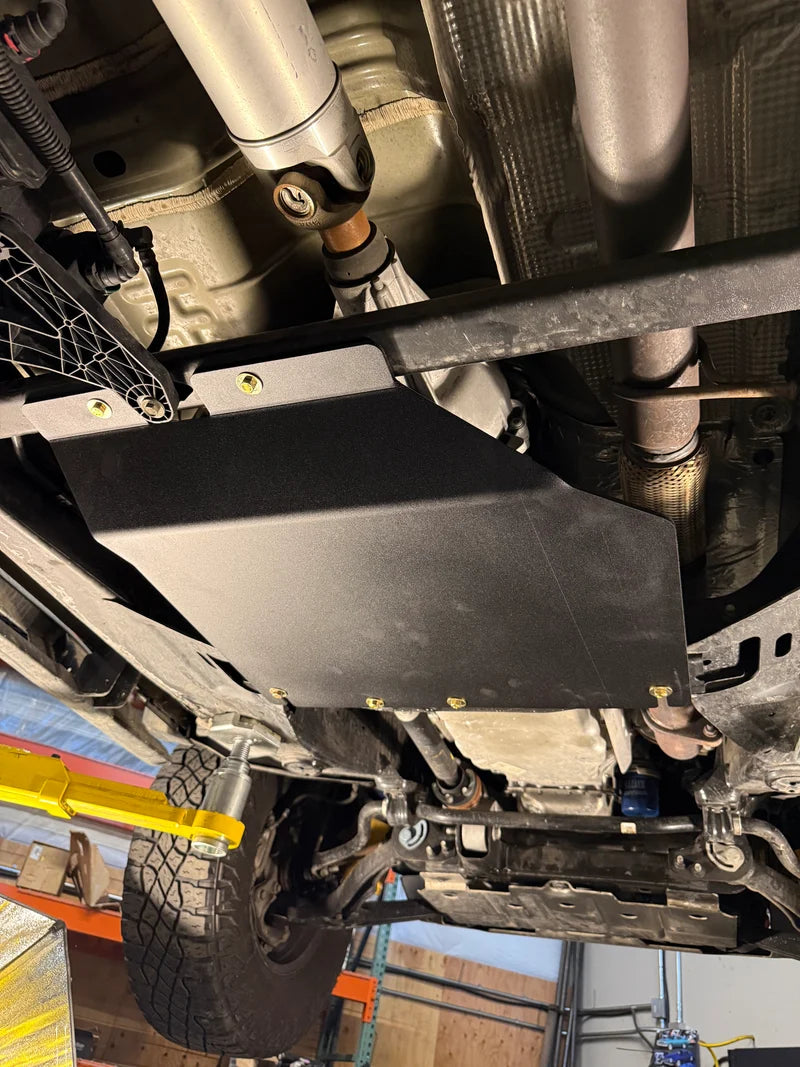 A black metal undercarriage splash shield plate mounted on a vehicle, shown from a low angle inside a garage or workshop with a tire, suspension components, and exhaust visible.