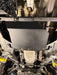 A black metal undercarriage splash guard plate mounted on a vehicle's chassis, secured with multiple gold-colored bolts. The background shows various automotive undercarriage components, pipes, and mechanical parts.