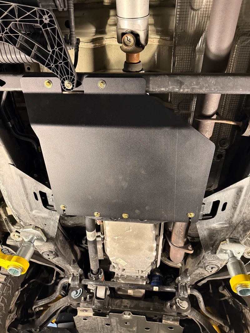 A black metal undercarriage splash guard plate mounted on a vehicle's chassis, secured with multiple gold-colored bolts. The background shows various automotive undercarriage components, pipes, and mechanical parts.