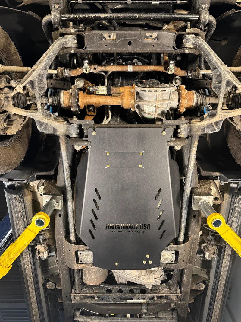 A black steel underbody shield with the text 'Jubereen Auto' printed on it, installed on the bottom of a vehicle. The vehicle is shown underneath on a lift, with yellow wheel chocks visible on both sides.