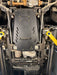 A black metal undercarriage splash shield with mounting holes and a printed logo, installed on a vehicle's chassis.
