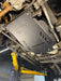 A black metal undercarriage splash shield with the text 'JAGUARHAUT USA' printed on it, installed under a vehicle on a yellow hydraulic lift in an auto repair shop.