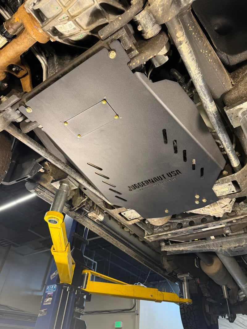 A black metal undercarriage splash shield with the text 'JAGUARHAUT USA' printed on it, installed under a vehicle on a yellow hydraulic lift in an auto repair shop.