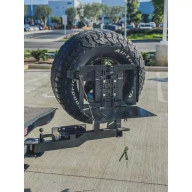 Rigid Armor Swing out Tire carrier system with a tire on a Land Cruiser, with a blurred background of trees and buildings.