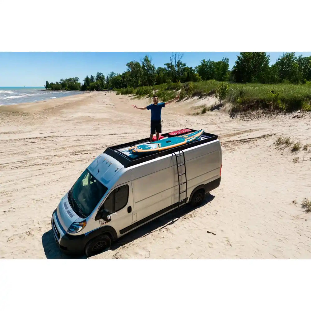 Van with a surfboard on top parked on a sandy beach with a person standing nearby.