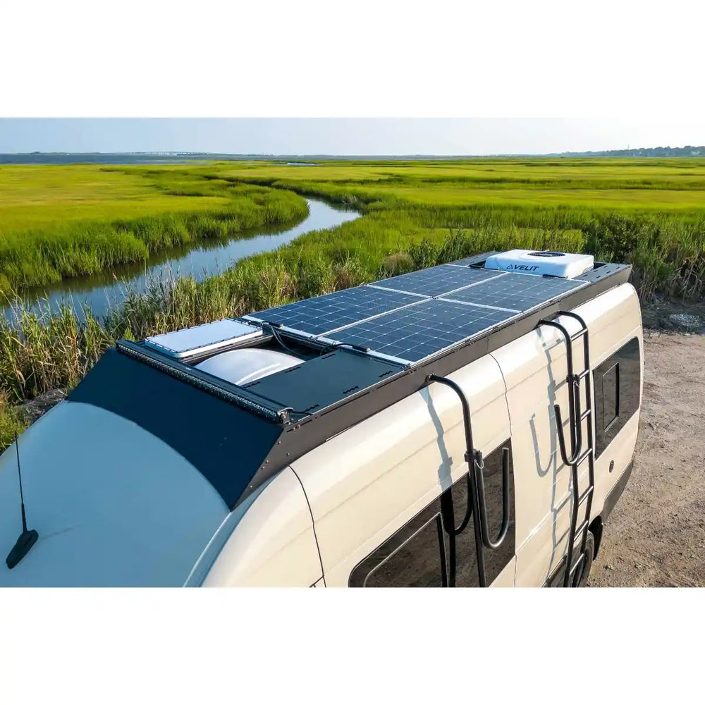 Sprinter with solar panels on the Orion roof rack parked near a body of water and greenery