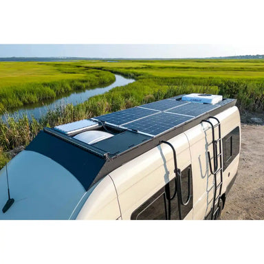 Sprinter with solar panels on the Orion roof rack parked near a body of water and greenery
