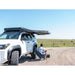 Person adjusting gear beside white 4Runner on gravel, black RCI roof rack with awning extended