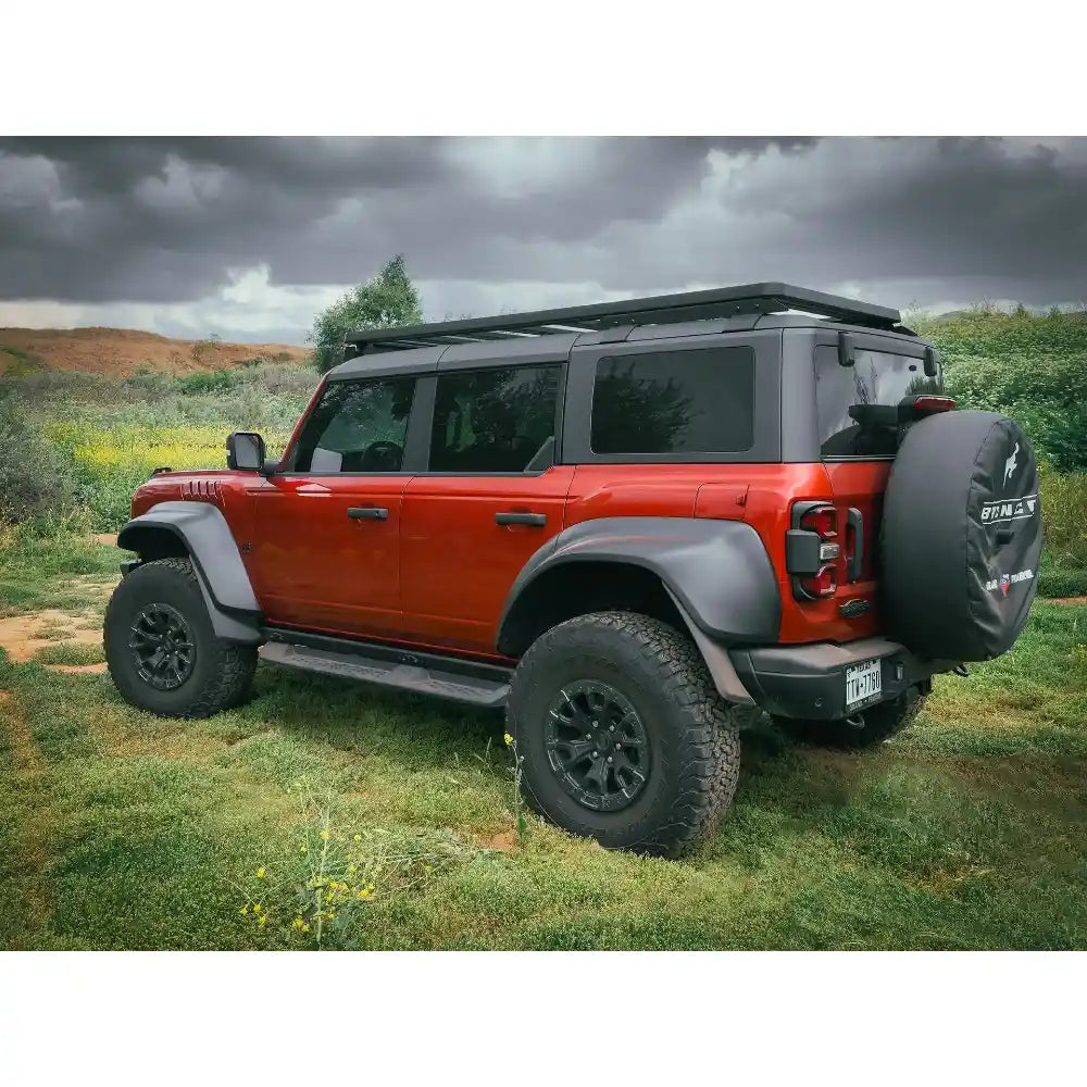 Side view of Red and gray off-road Bronco with roof rack on a grassy field under a cloudy sky.