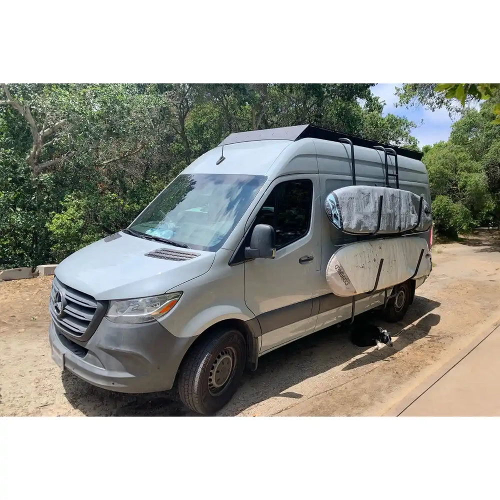 Silver van with a roof rack and surfboard on a road surrounded by trees