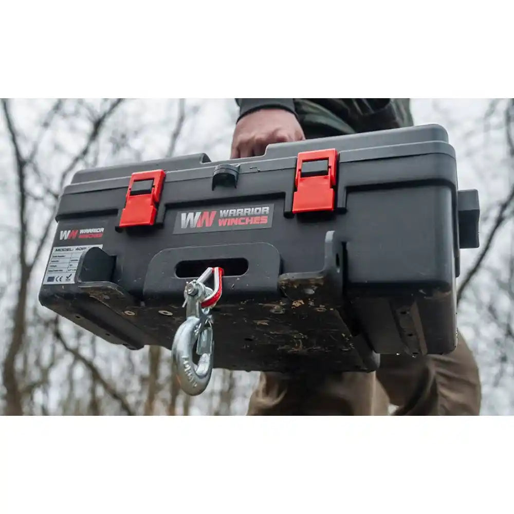 Person carrying a large gray Trojan 12v Portable Utility Winch with red latches on a blurred outdoor background