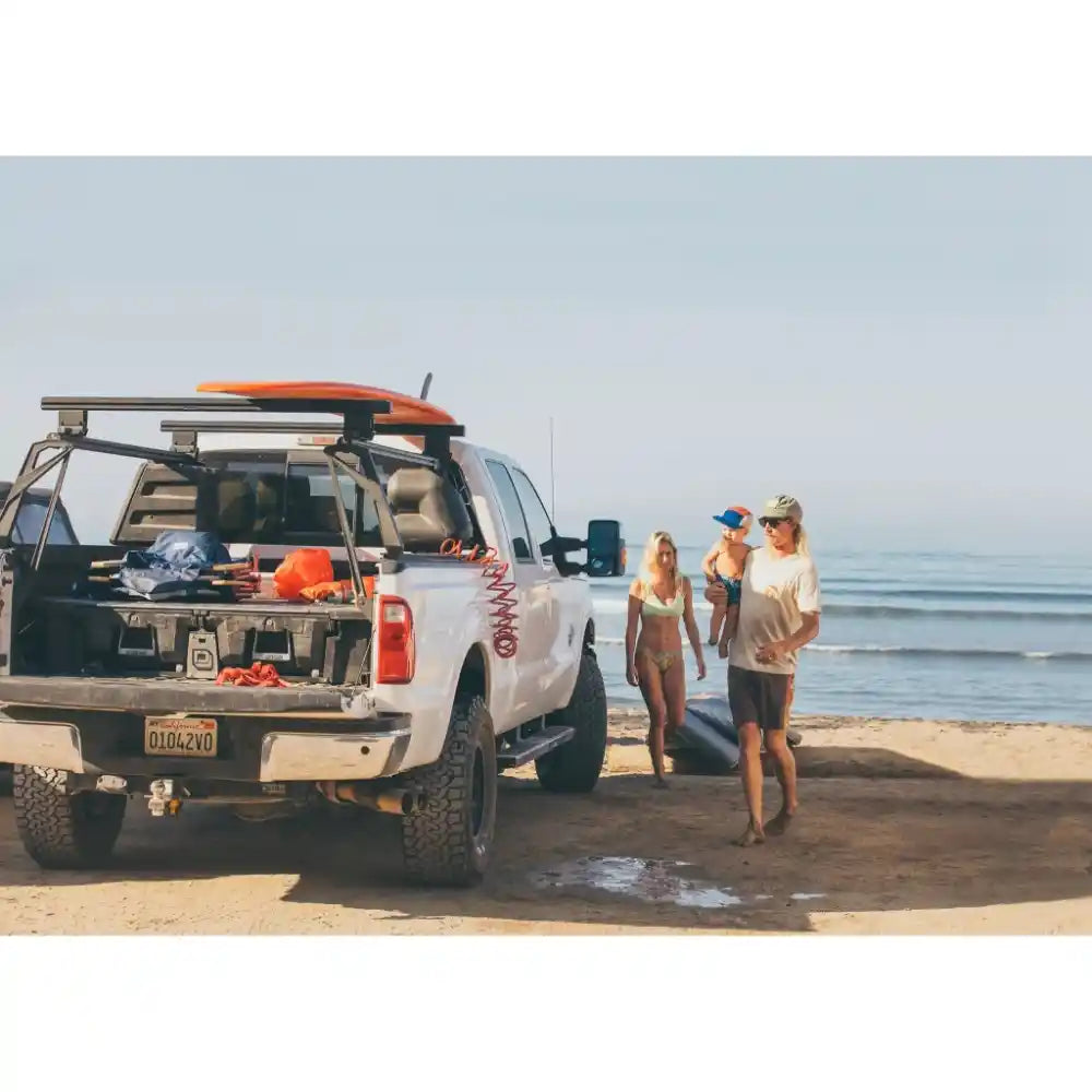 White Ford truck with surfboard in a bed rack on a beach, people standing nearby