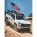 White GMC truck on a dirt road with installed Active Cargo System Bed Rack by Leitner Designs and an American flag on top, against a clear blue sky.