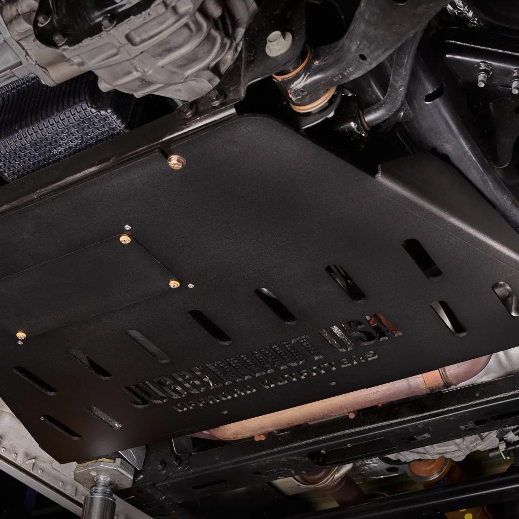 Close-up underbody view of the transmission skid plate installed near the catalytic converter and exhaust, mounted with visible bolts