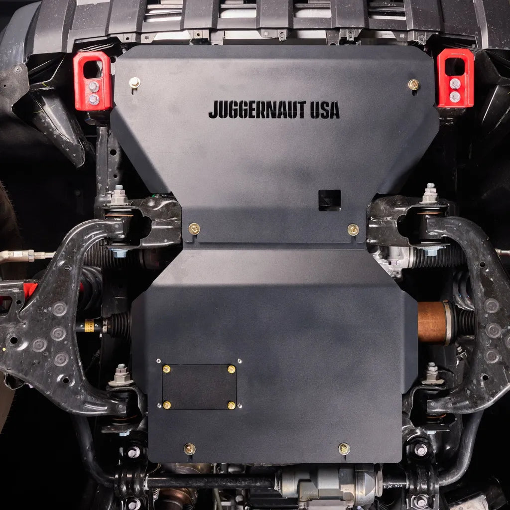 Straight-on underbody view of the engine skid plate installed, showing the JUGGERNAUT USA logo and red-capped mounting hardware