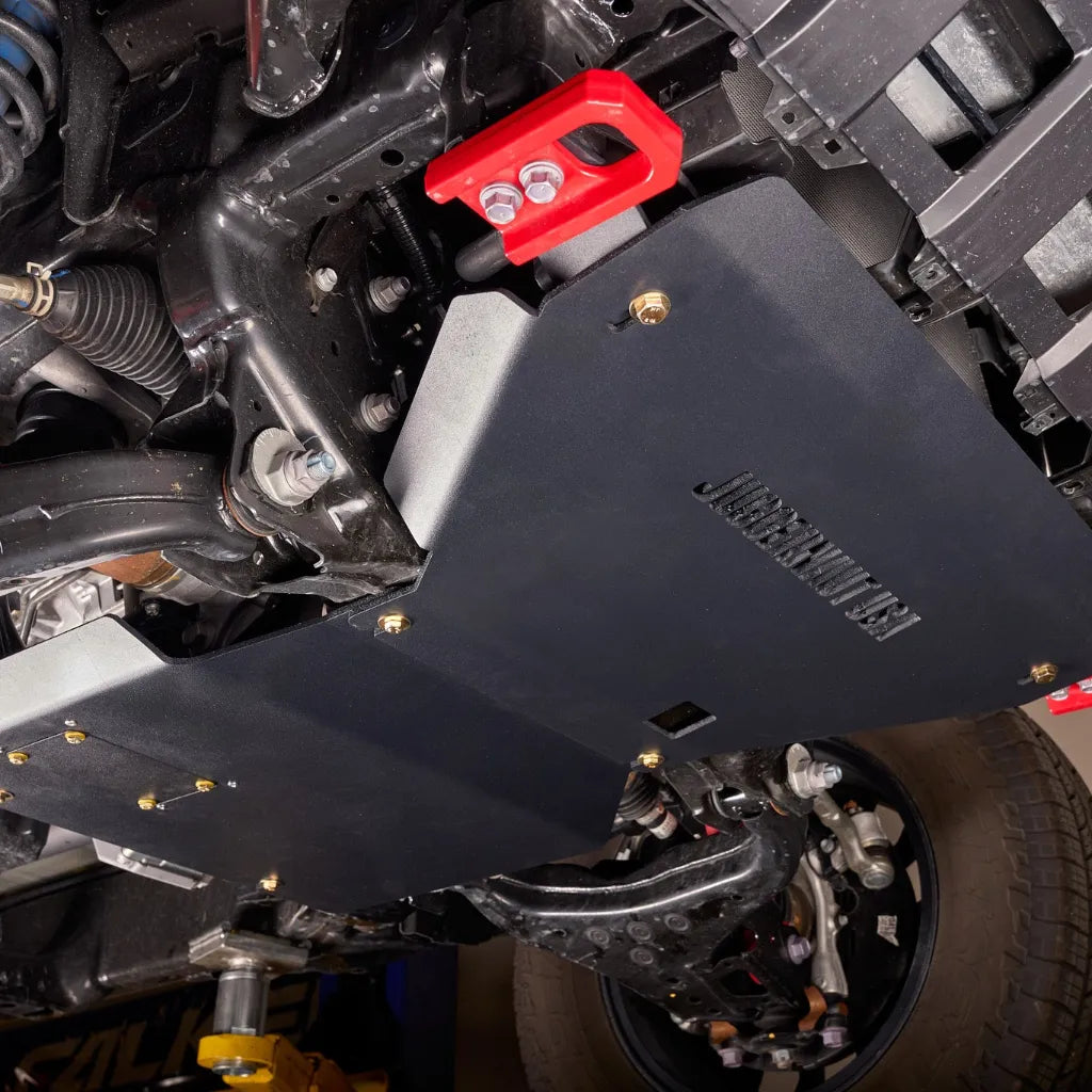 Underbody angle view of the engine skid plate installed near the front wheel, showing red-capped fasteners