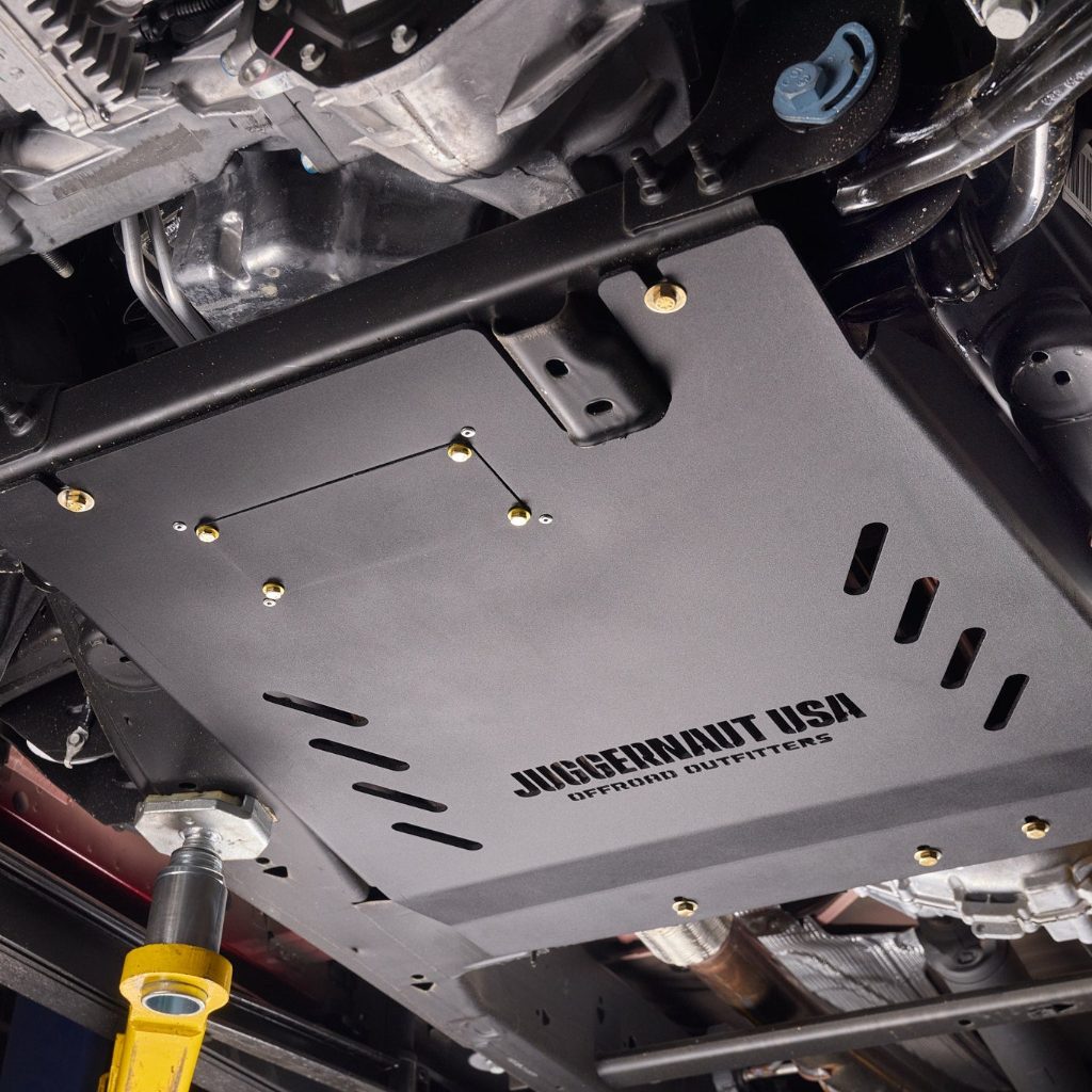 Angled underbody view of the Juggernaut USA Silverado 1500 transmission skid plate, showing the diagonal drainage slots and mounting hardware along the frame rail