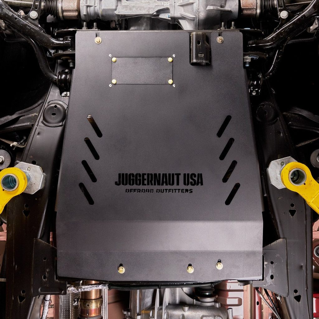 Juggernaut USA transmission skid plate and catalytic converter shield installed on a Chevy Silverado 1500, straight-on underbody view showing the removable oil-drain access panel and mounting bolts