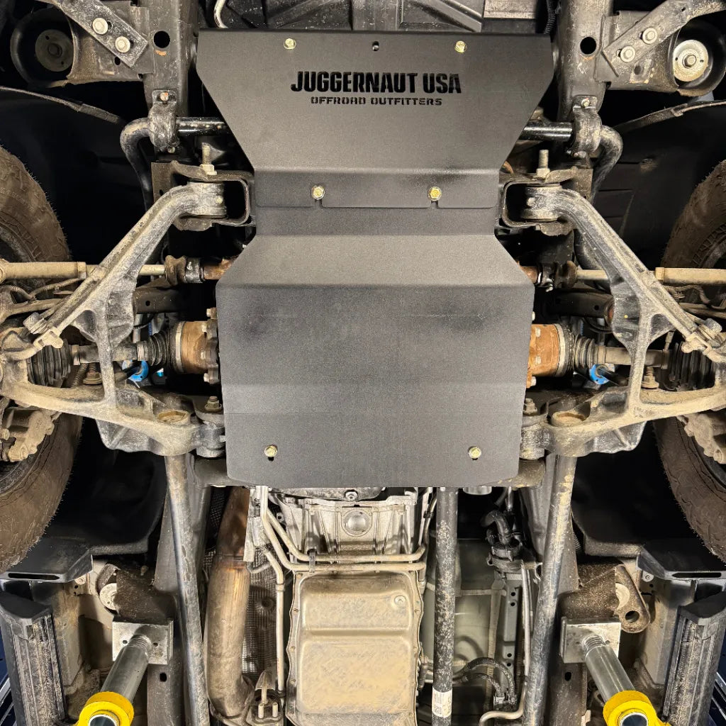 Wide underbody view of the engine skid plate centered between both front wheels, with the transmission visible just behind it