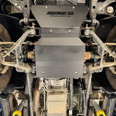 Wide underbody view of the engine skid plate centered between both front wheels, with the transmission visible just behind it