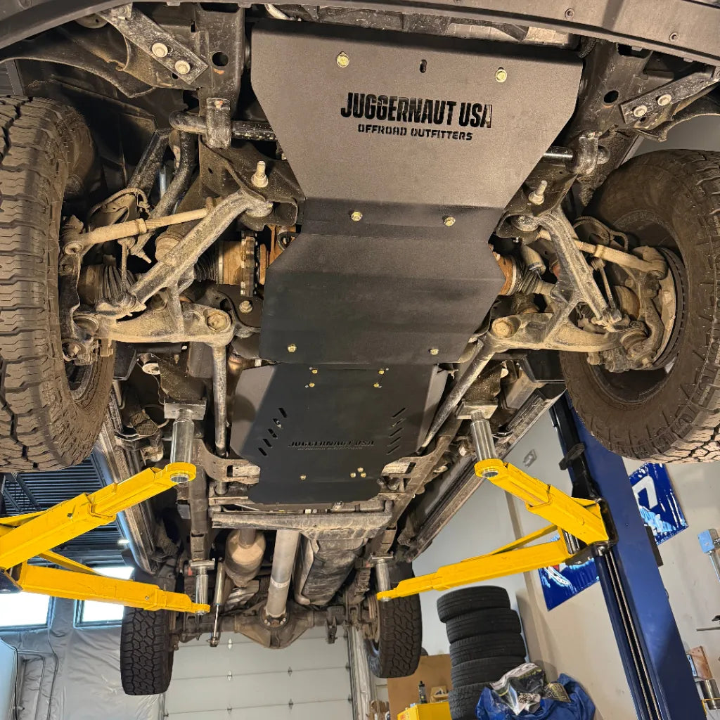 Front-quarter underbody view of the complete Juggernaut USA Silverado HD skid plate package on a shop lift, both front wheels visible