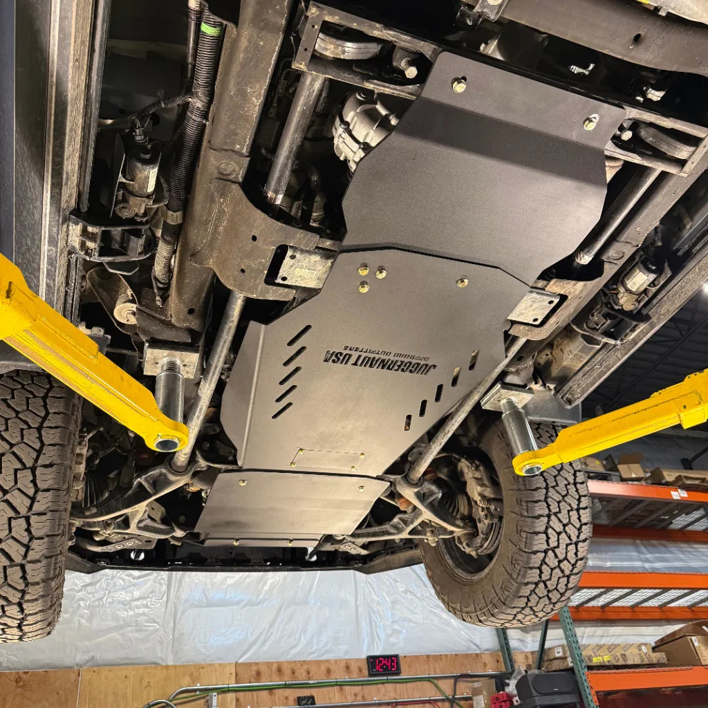 Angled underbody view of the transmission and transfer case skid plate installed in a home garage, showing drainage slots and mounting hardware
