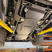 Angled underbody view of the transmission and transfer case skid plate installed in a home garage, showing drainage slots and mounting hardware