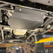 Front-quarter underbody view of the transfer case skid plate on a shop lift, with the driver-side wheel and tire visible