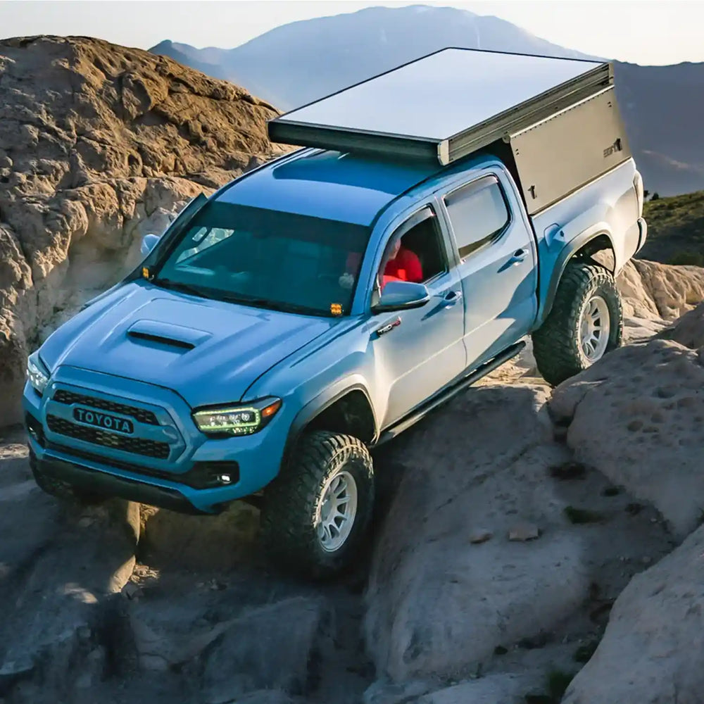 Blue Toyota Tacoma with rock sliders and a camper shell driving on rocky terrain