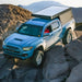 Blue Toyota Tacoma with rock sliders and a camper shell driving on rocky terrain