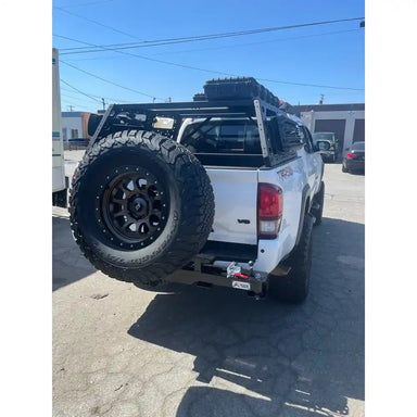 White Tacoma with large black tire mounted on Rigid Armor Swing Out Tire Carrier and off-road accessories in a parking lot.