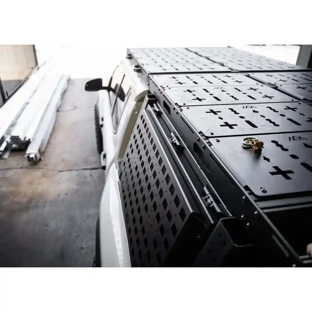 Black metal storage rack attached to a white vehicle with tools on top, on a concrete surface.